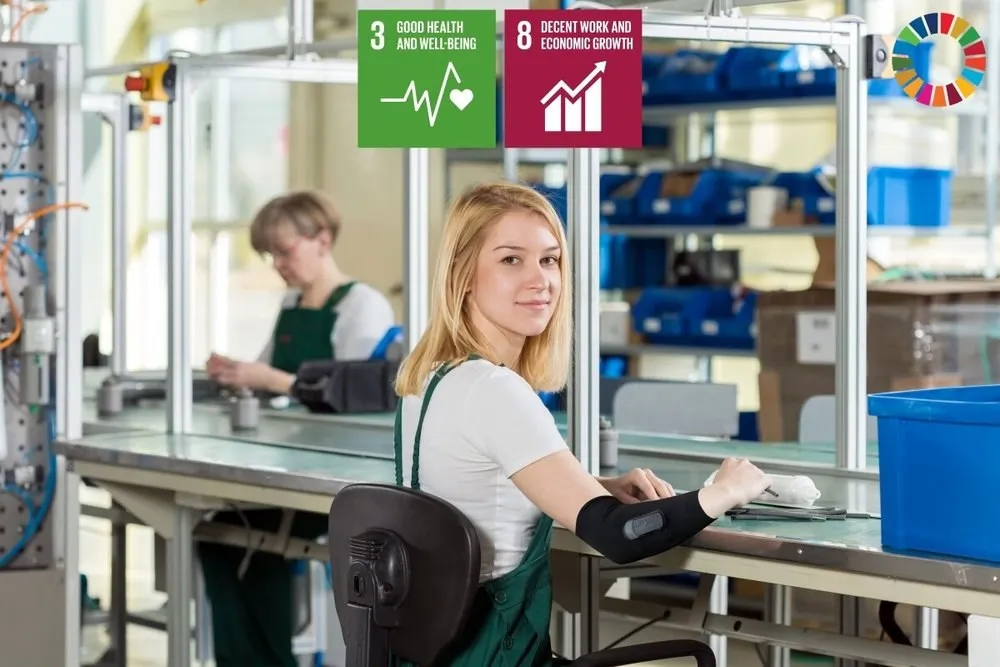 Factory worker at a workbench wearing an elbow sensor sleeve, with the icons for UN goals 3 and 8