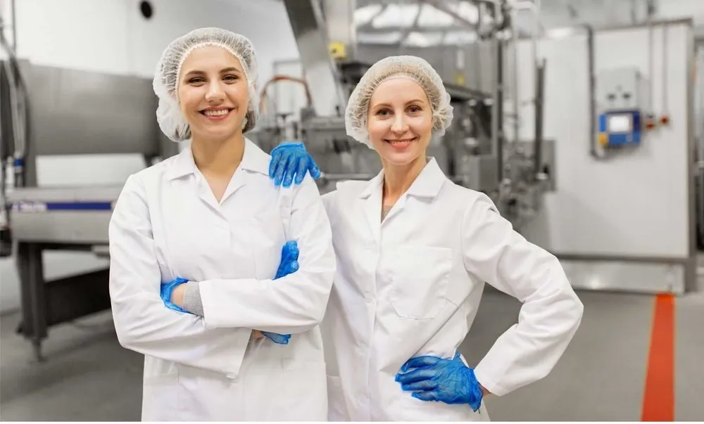 Two food-industry workers in lab coats, hairnets and blue gloves in a production hall